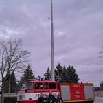 Sirene hoch auf Metallpfosten dazu rotes Feuerwehrauto und 5 Kameraden der Feuerwehr