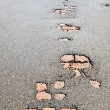 schadhafte Asphaltdecke mit Löchern, durch welche bereits das darunterliegende Kopfsteinpflaster sichtbar wird