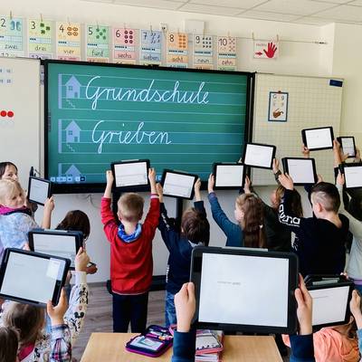 Kinder stehen in einem Klassenraum vor einer digitalen Tafel und halten Tablets in die Höhe